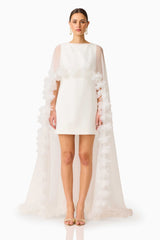 Model wearing the Ellie Cape Mini Dress in White with floor length cape with floral trim front shot