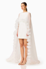 Model wearing the Ellie Cape Mini Dress in White with floor length cape with floral trim side shot