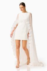 Model wearing the Ellie Cape Mini Dress in White with floor length cape with floral trim posed walking shot