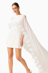 Model wearing the Ellie Cape Mini Dress in White with floor length cape with floral trim close up walking shot
