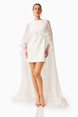 Model wearing the Ellie Cape Mini Dress in White with floor length cape with floral trim posed hand on hips shot